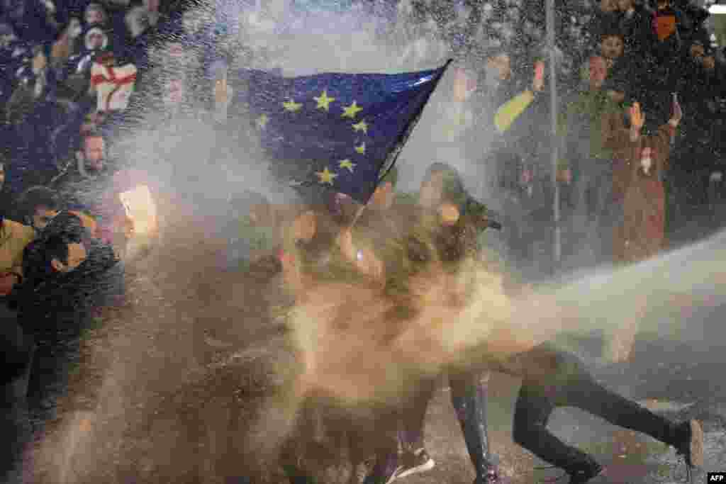 Протестующие, размахивающие флагом Европейского Союза, под огнем водометов во время столкновений с полицией возле парламента Грузии в Тбилиси, 7 марта 2023 года. Грузинская полиция применила слезоточивый газ и водометы против протестующих. Люди выступали против спорного законопроекта об "иностранных агентах". (Photo by AFP)